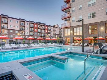 A swimming pool surrounded by lounge chairs and umbrellas in front of apartment buildings.